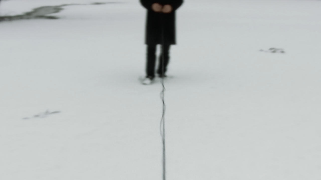 Un homme - dont on ne voit que le bas du corps et pas le visage - tient un fil qui s'étend jusqu'au premier plan de l'image, sur un fond de neige.