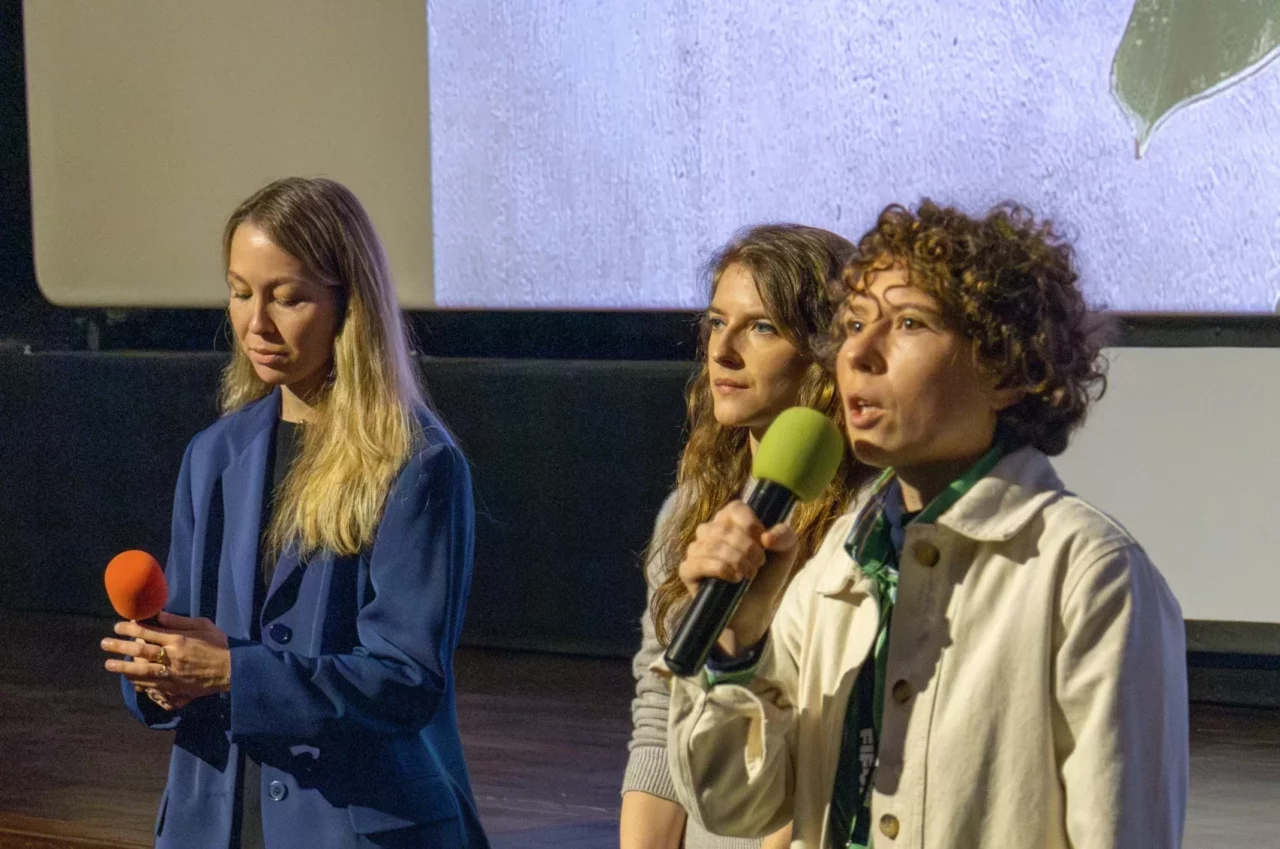 Trois femmes vues de près, l'une tient un micro et s'adresse au public